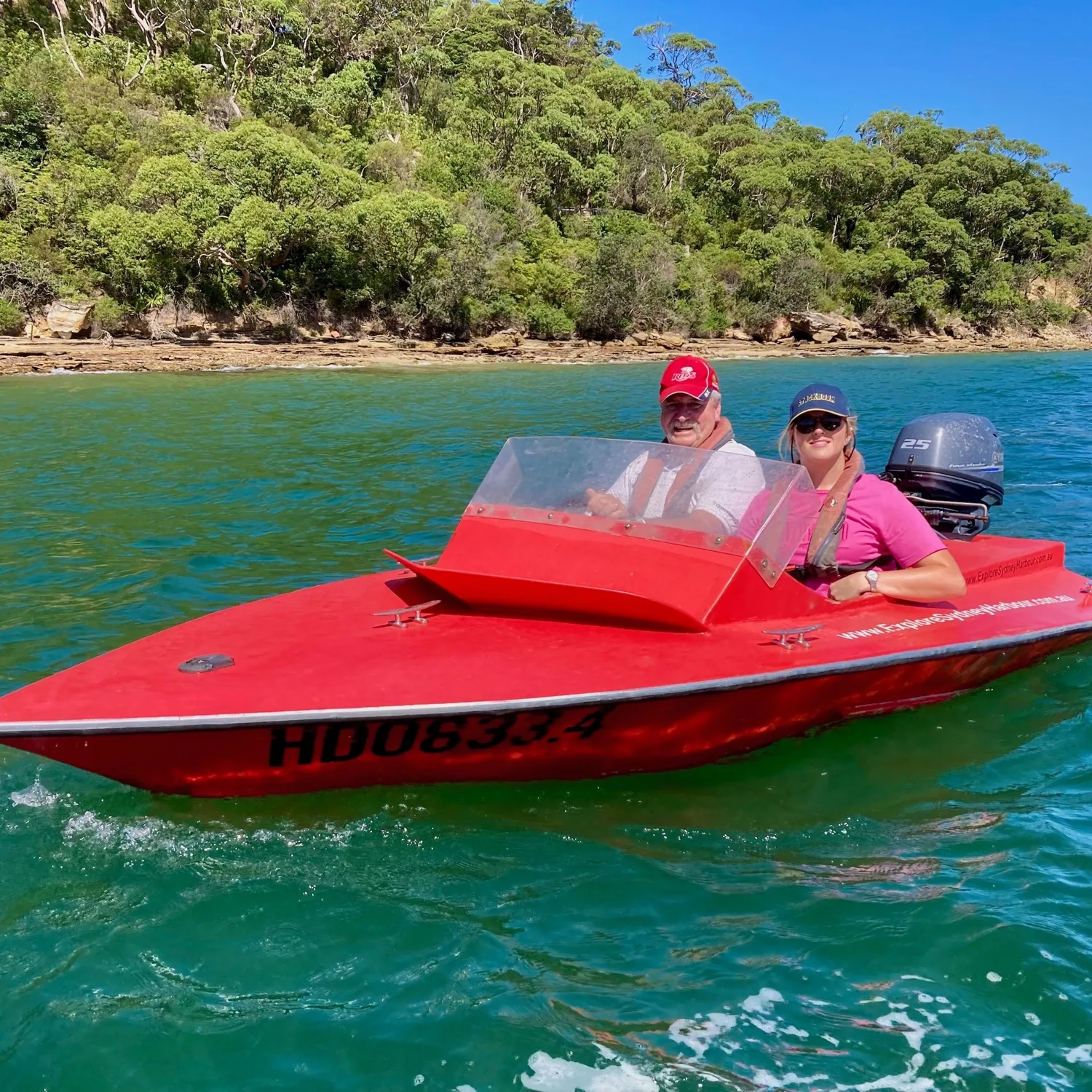 Two guests driving a red speedboat past bushland on Sydney Harbour